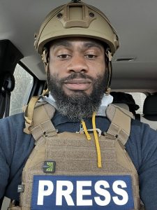 Man sitting in a car wearing body armor that has the word "Press" issued on the front.